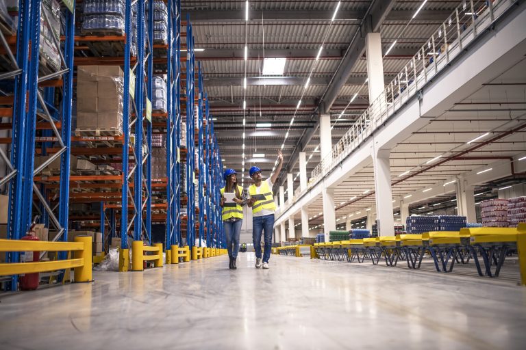 distribution-warehouse-interior-with-workers-wearing-hardhats-reflective-jackets-walking-storage-area-768x512